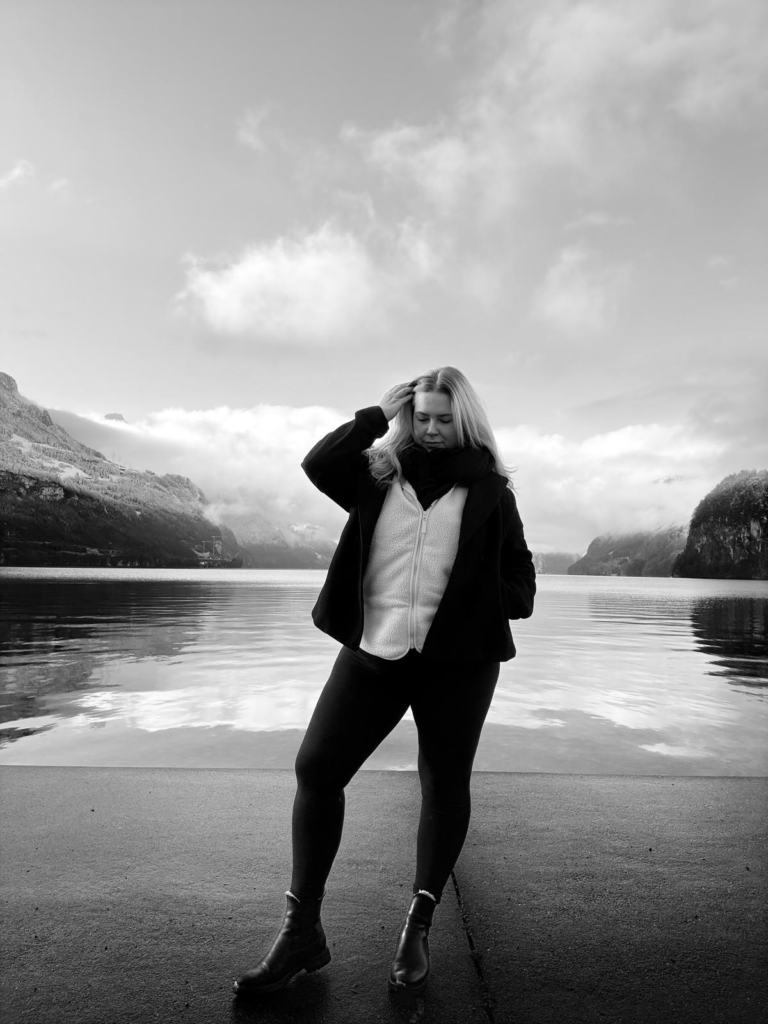 Schwarz weiss Bild einer Frau, die nach unten auf den Boden blickt, hinter ihr der See und Berge