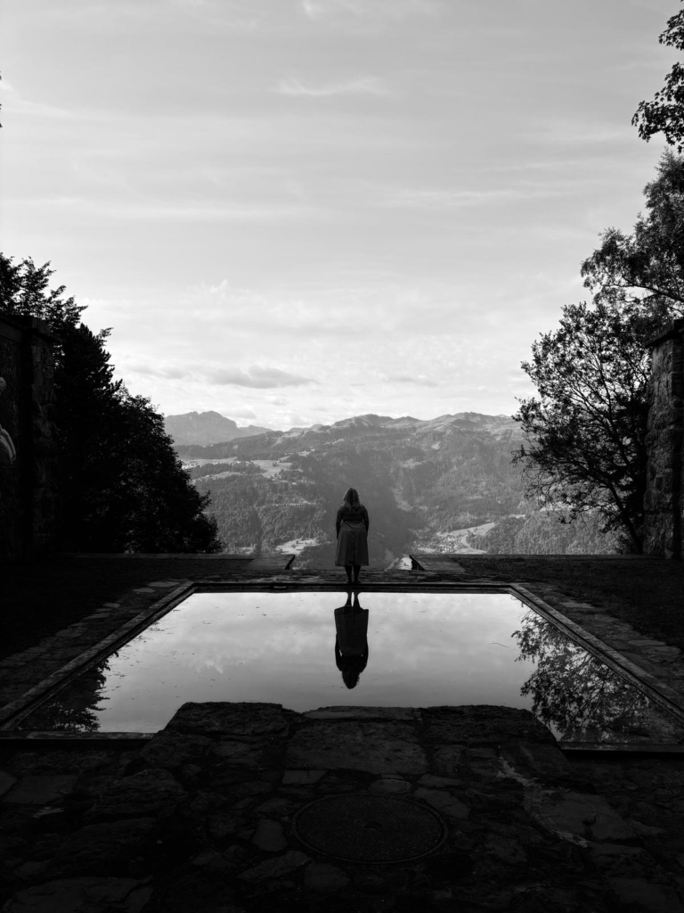 Schwarz weiss Bild von einer Frau, die über den Bergen steht hinter hier ist Wasser und sie Spiegelt sich darin